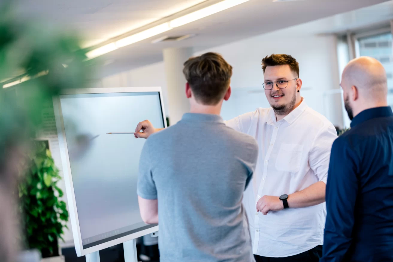 Geschäftsmann zeigt vor Kollegen auf Flipchart in grünem Bürobereich.