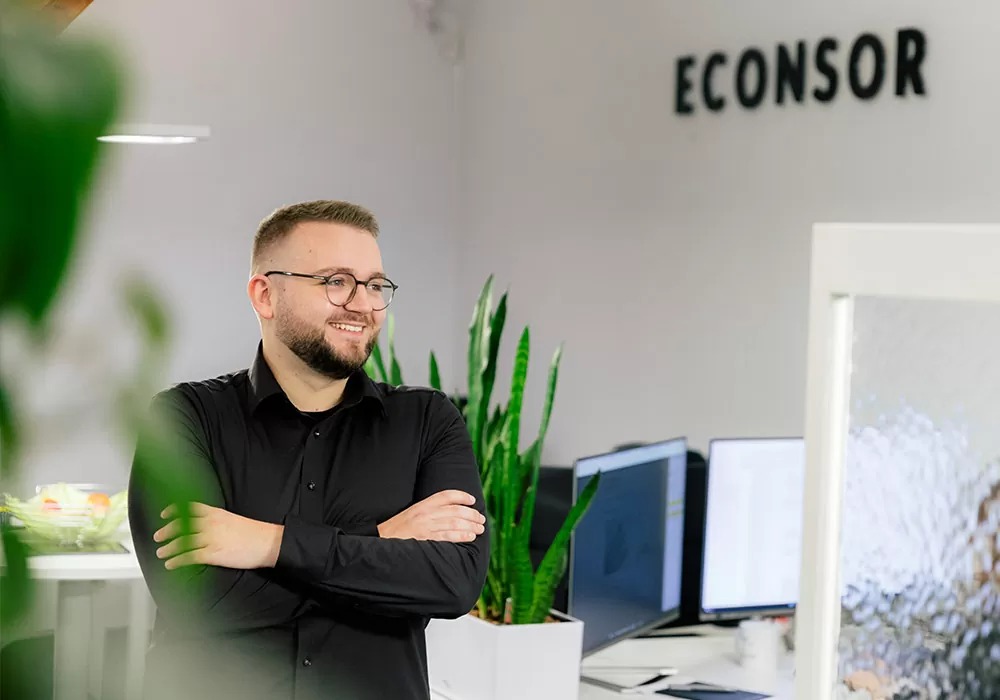 Junger mann steht in einem Büro. Im Hintergrund sieht man Arbeitsplätze.