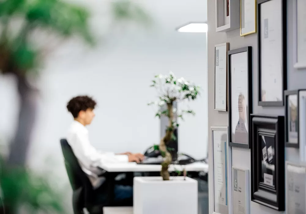 Blick in ein modernes Büro. Rechts an der Wand hängen Auszeichnungen. Im Hintergrund sieht man einen jungen Mann am Arbeitsplatz vor einen PC sitzen.