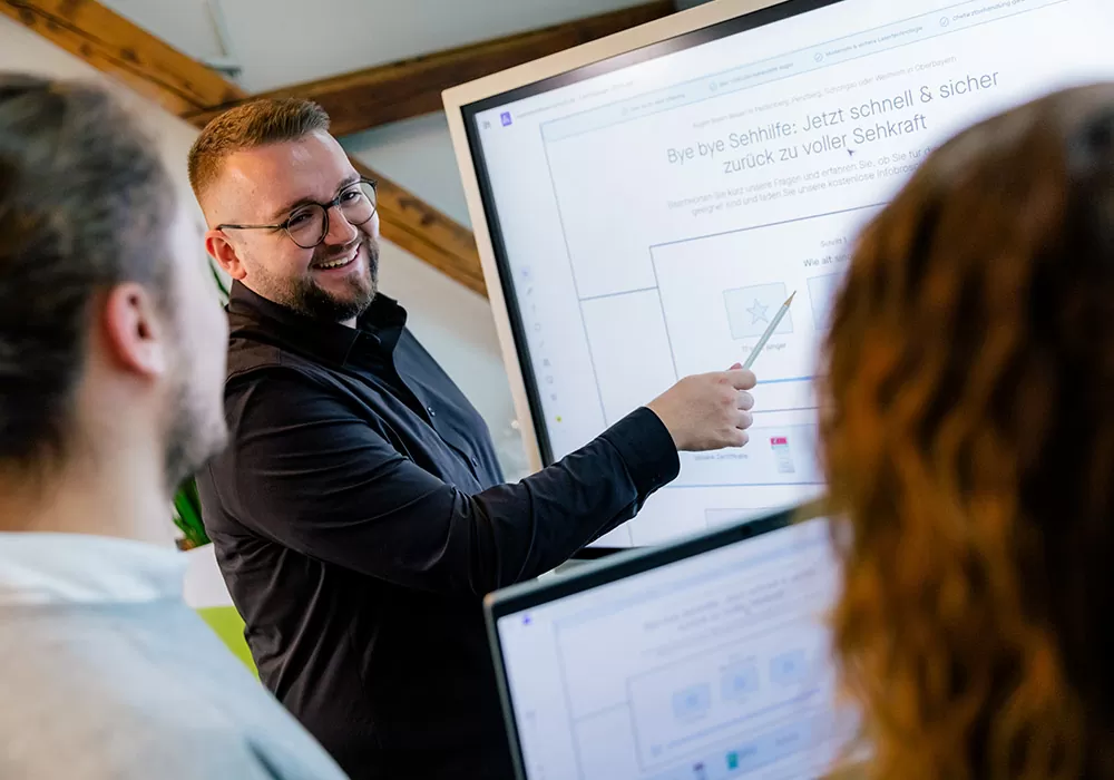 Junger Mann steht lächelnd vor einem Smartboard und zeigt darauf. Im Vordergrund sieht man die Silhouetten zweier weiterer Personen.