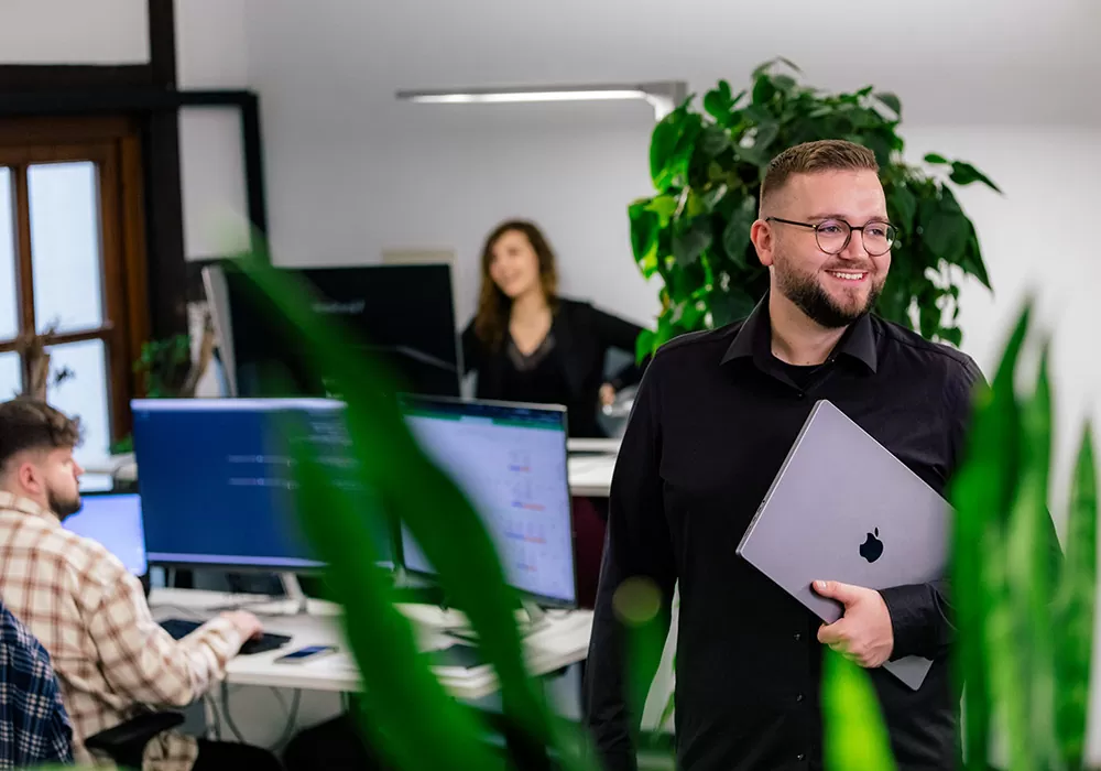 Mann mit Laptop im Arm im Vordergrund. Im Hintergrund sieht man zwei weitere Menschen die an einem Arbeitsplatz sitzen und arbeiten.