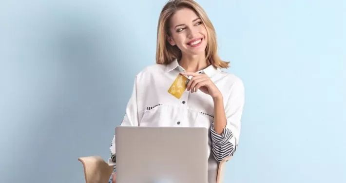 Eine Frau sitzt vor ihrem Laptop mit einer Kreditkarte in der Hand und lächelt.
